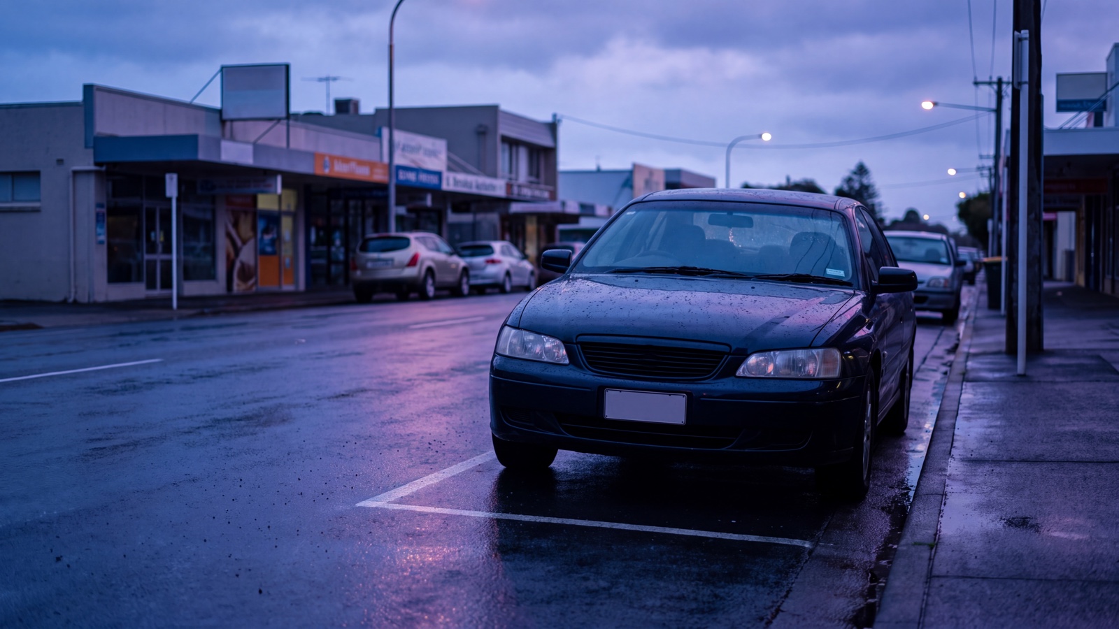 Generic coastal service-centre street after rain, with an ordinary parked vehicle and restrained cobalt and purple reflections.