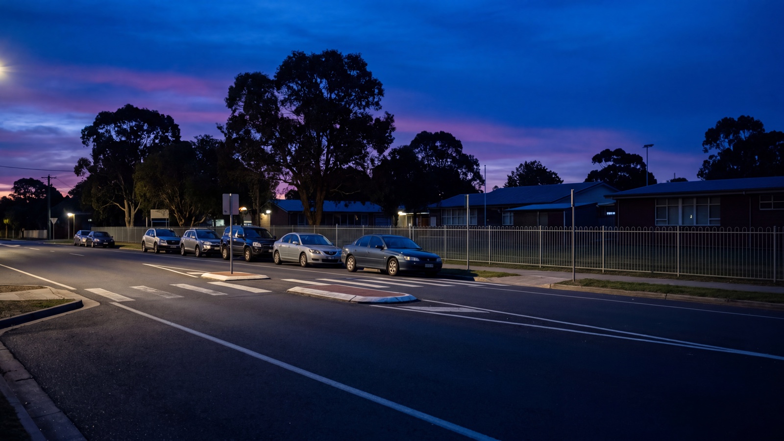 Generic school-frontage road outside active pick-up time, with ordinary parked vehicles, a plain fence and mature trees at blue hour.