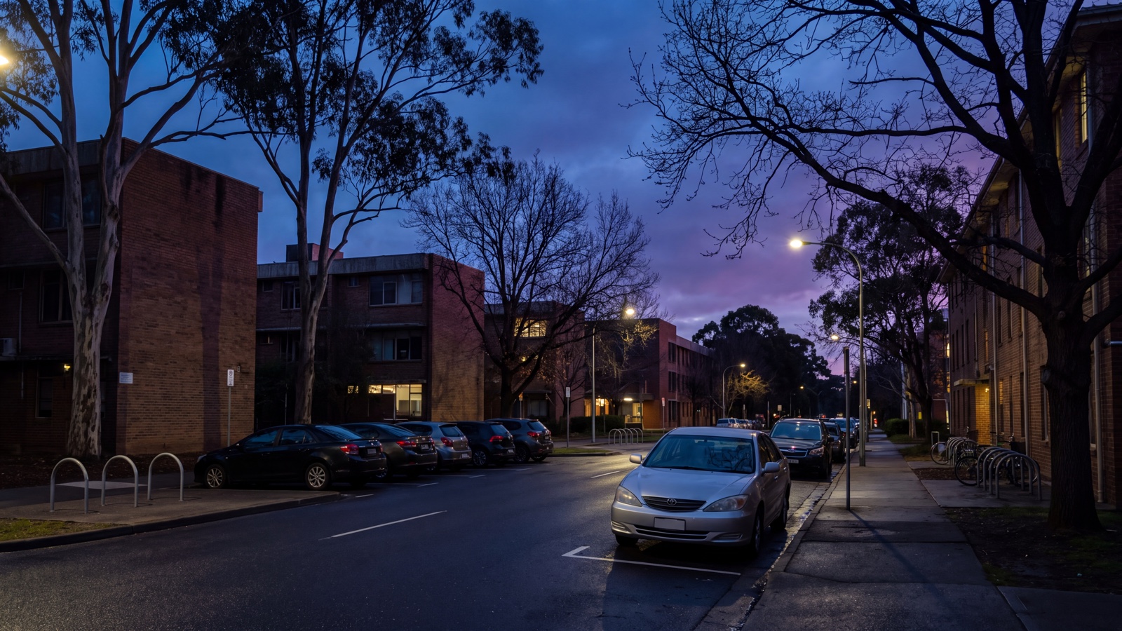 Generic university-precinct edge street with parked vehicles, bicycle racks and non-distinct academic buildings at blue hour.